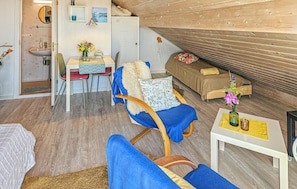 A cozy room with a wooden ceiling, a dining area with a table and chairs, a sofa with a floral cushion, and a small table with a vase of flowers.