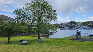 A grassy park area with a large tree, a playground, and a view of a marina with boats.