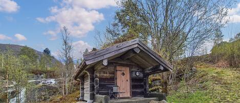 Una pequeña cabaña de madera con un porche, rodeada de árboles y un paisaje montañoso.