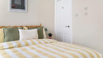 Um quarto com cama, almofadas, mesa de cabeceira, porta e um objeto fixado na parede.