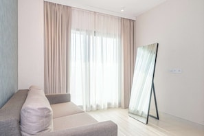 Un salon moderne avec un canapé, de grandes fenêtres ornées de rideaux légers et un grand miroir sur toute la hauteur du mur.