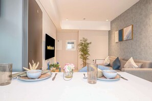A modern living room with a sofa, a dining table set with bowls and cutlery, a television, and a large plant. - Longevity Superior Escape Tranquil Wellness (Jumeirah Village Triangle)