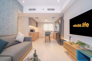 A modern living room with a sofa, a TV mounted on the wall, and a kitchen area in the background. - Longevity Superior Escape Tranquil Wellness (Jumeirah Village Triangle)