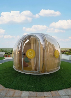 Une structure transparente en forme de dôme, dotée d’une porte, qui se trouve sur une pelouse verdoyante, avec un ciel dégagé en arrière-plan.
