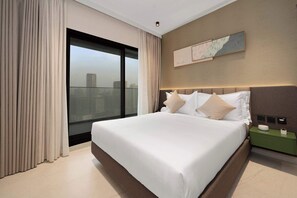 A hotel room with a large bed, a nightstand, a wall-mounted artwork, and a view of the city through a large window. - The Longevity Retreat | Luxury Wellness Stay (Jumeirah Village Triangle)