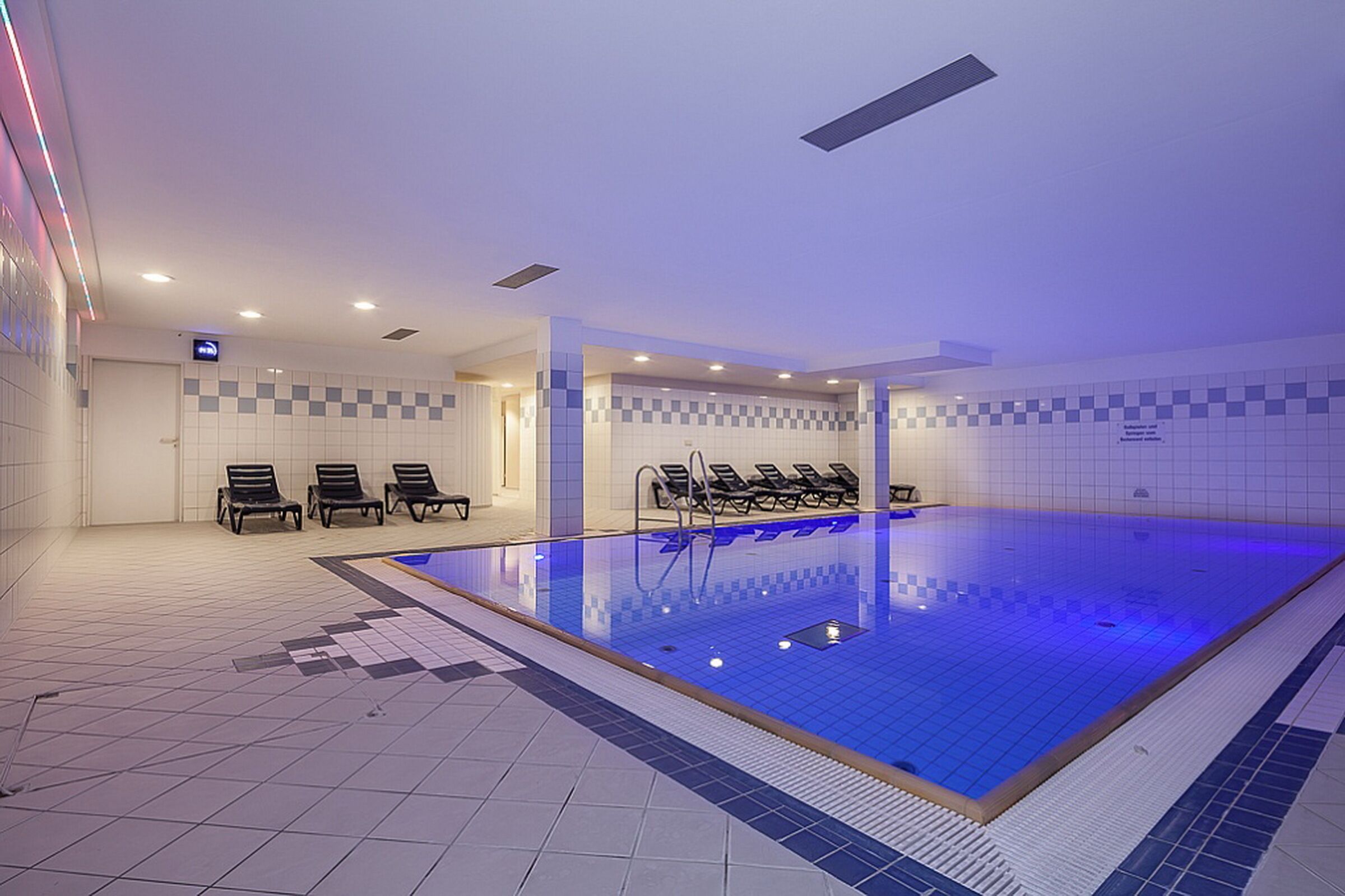 Une piscine intérieure avec des murs carrelés, un fond de piscine bleu et plusieurs chaises longues disposées autour de la piscine.