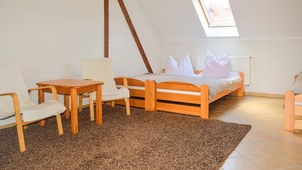 Un dormitorio con techo inclinado, una cama con ropa blanca, una mesa de madera, dos sillas y una alfombra marrón.