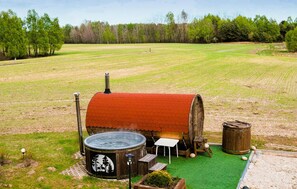 A hot tub with a wooden barrel exterior, a small table, and a wooden stool in a grassy field with trees in the background.