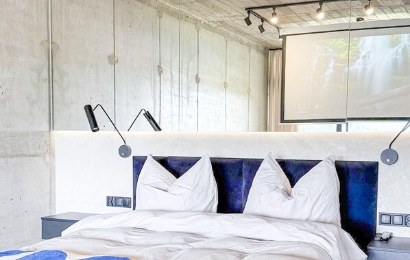 A modern bedroom with a large bed, a nightstand, a wall-mounted lamp, and a projector screen displaying a waterfall.