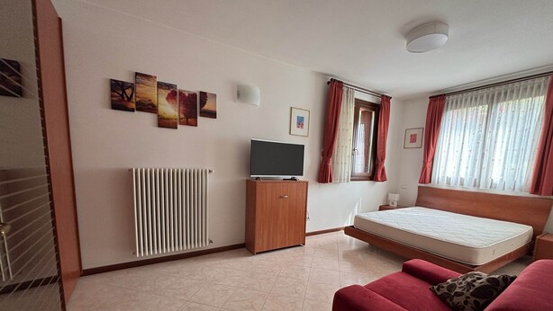 A hotel room with a bed, a TV, a radiator, a window with curtains, and artwork on the wall.