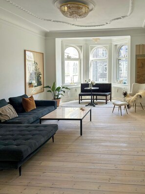 Ein geräumiges Wohnzimmer mit einem dunklen Sofa, einem Couchtisch und einem Sessel. Große Fenster sorgen für viel natürliches Licht.