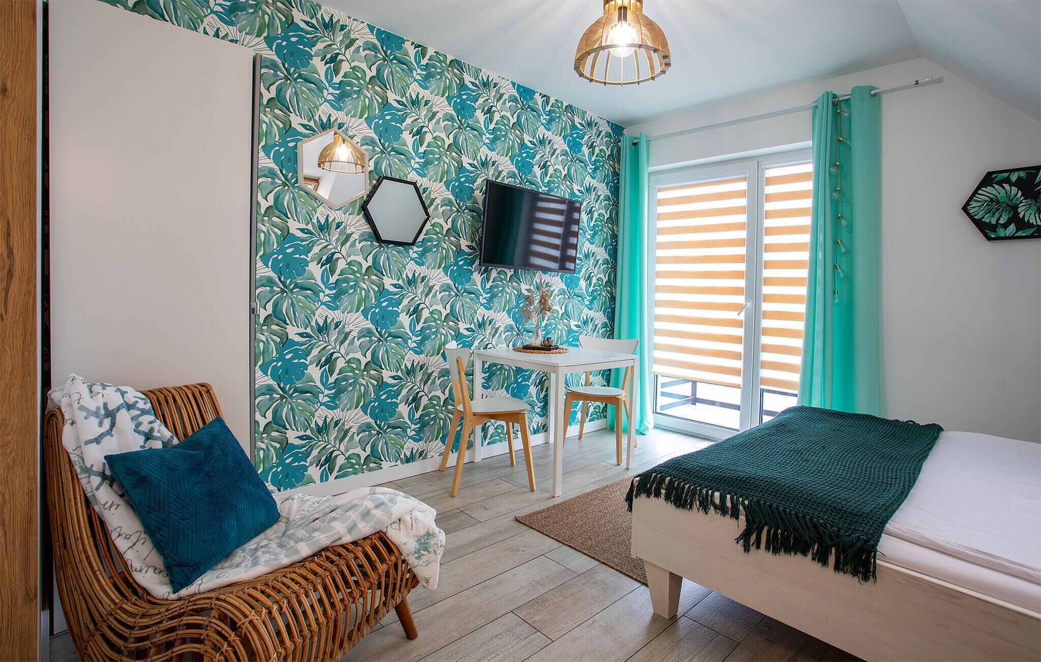 A room with a bed, a wicker chair, a small table with chairs, a TV, and a window with curtains.