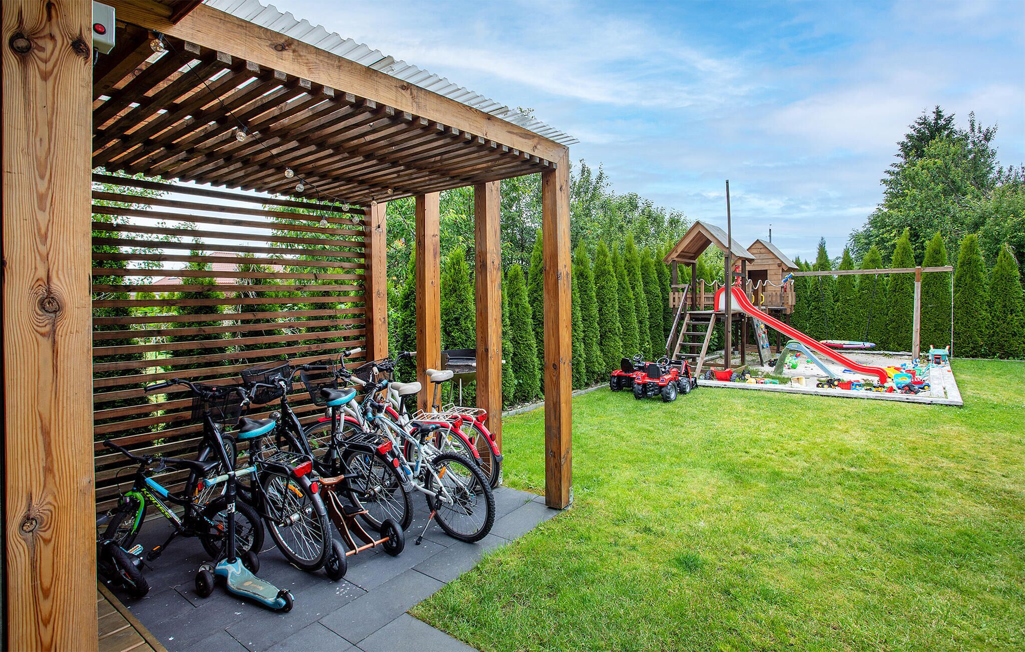 A covered bike parking area with bicycles, a playground with a slide, and a wooden playhouse.