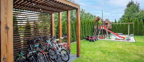 A covered bike parking area with bicycles, a playground with a slide, and a wooden playhouse.