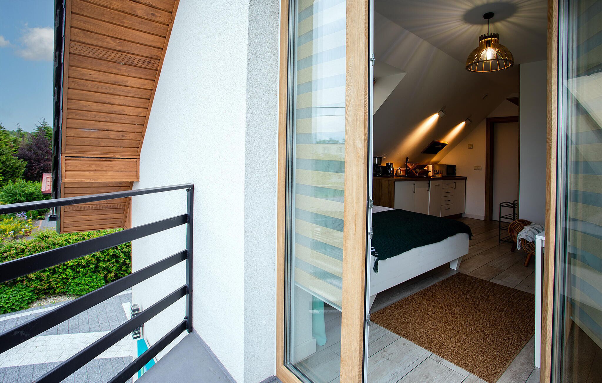 A modern bedroom with a glass door leading to a balcony, a bed with a green cover, and a kitchen area with a hanging light fixture.