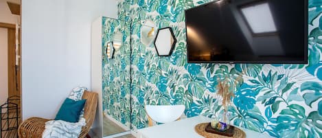 Una habitación con un televisor grande montado en la pared, una mesa blanca con un objeto decorativo, una silla de mimbre con un cojín azul y una pared con un diseño de hojas tropicales.