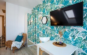 A room with a large TV mounted on the wall, a white table with a decorative item, a wicker chair with a blue cushion, and a wall with a tropical leaf pattern. - Gorgeous apartment in Nowecin with WiFi (Nowecin)
