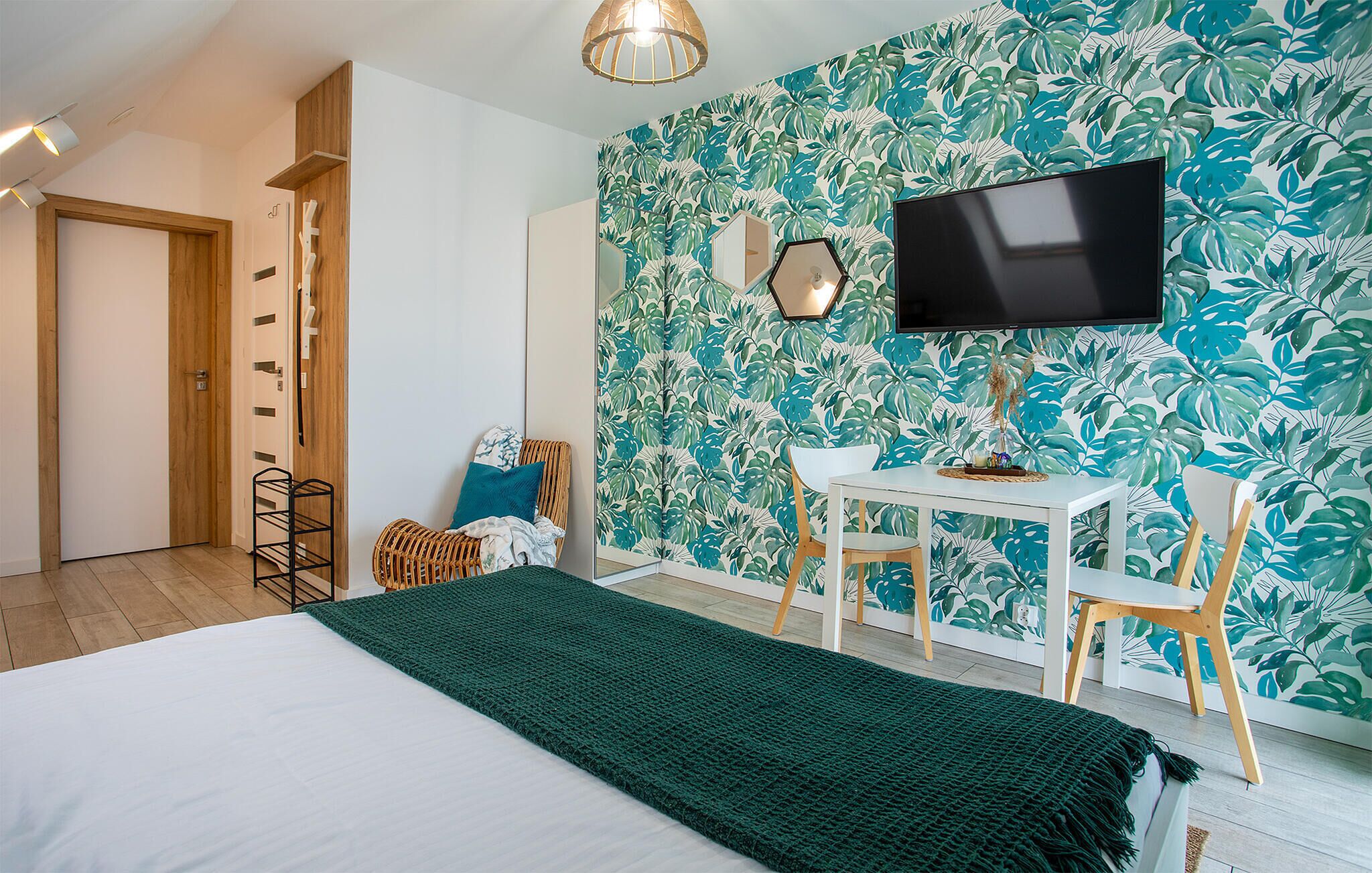 A room with a bed, a desk, chairs, a television, and a wall with a leafy pattern.
