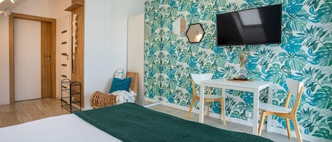 A room with a bed, a desk, chairs, a television, and a wall with a leafy pattern.