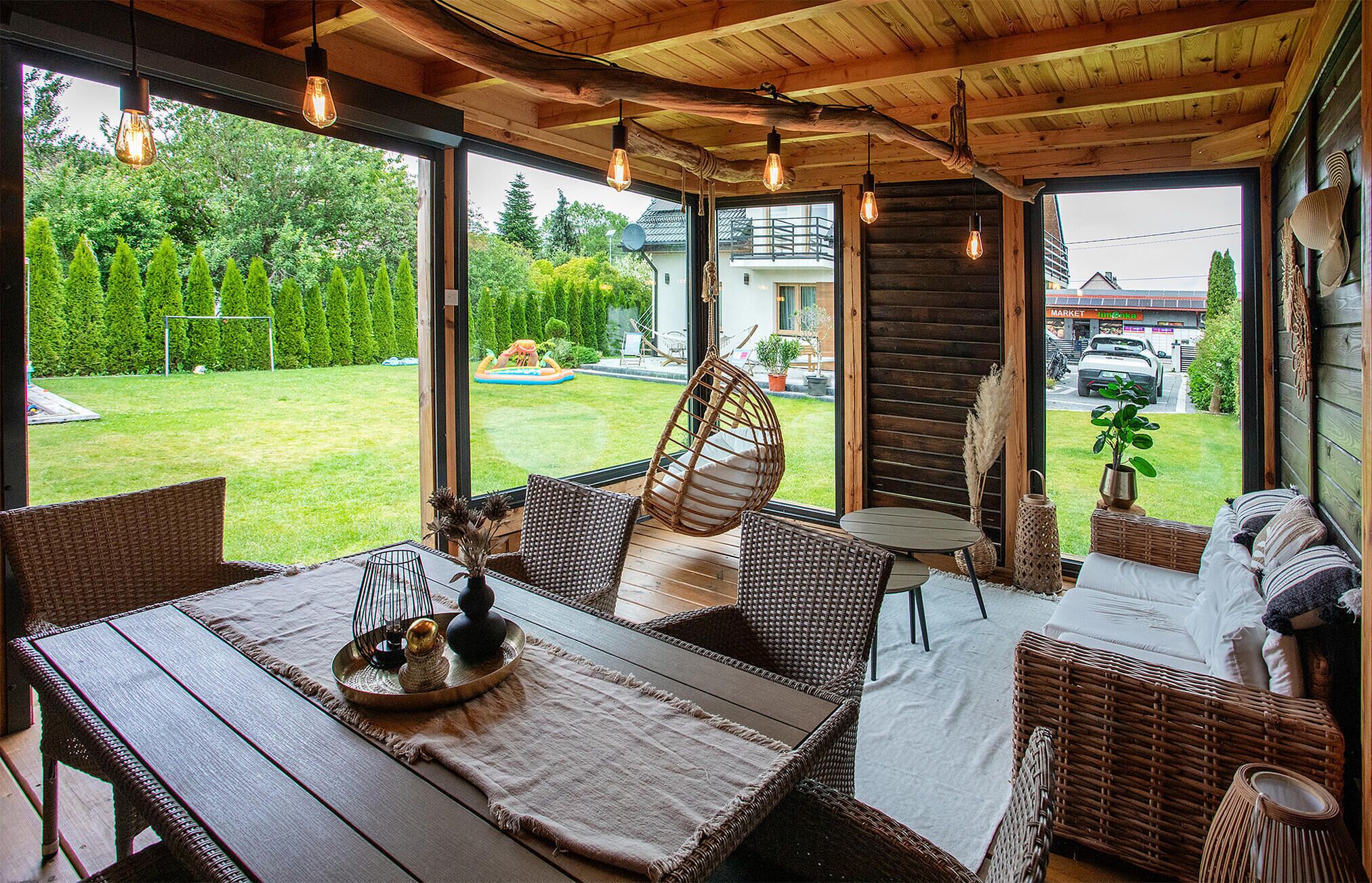 A cozy outdoor seating area with wicker furniture and a hanging chair, overlooking a well-maintained garden and a view of a building with a sign that reads "MARKET".