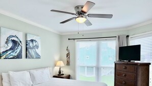 Un dormitorio con cama, ventilador de techo, obras de arte, mesita de noche con lámpara, cómoda y televisor.