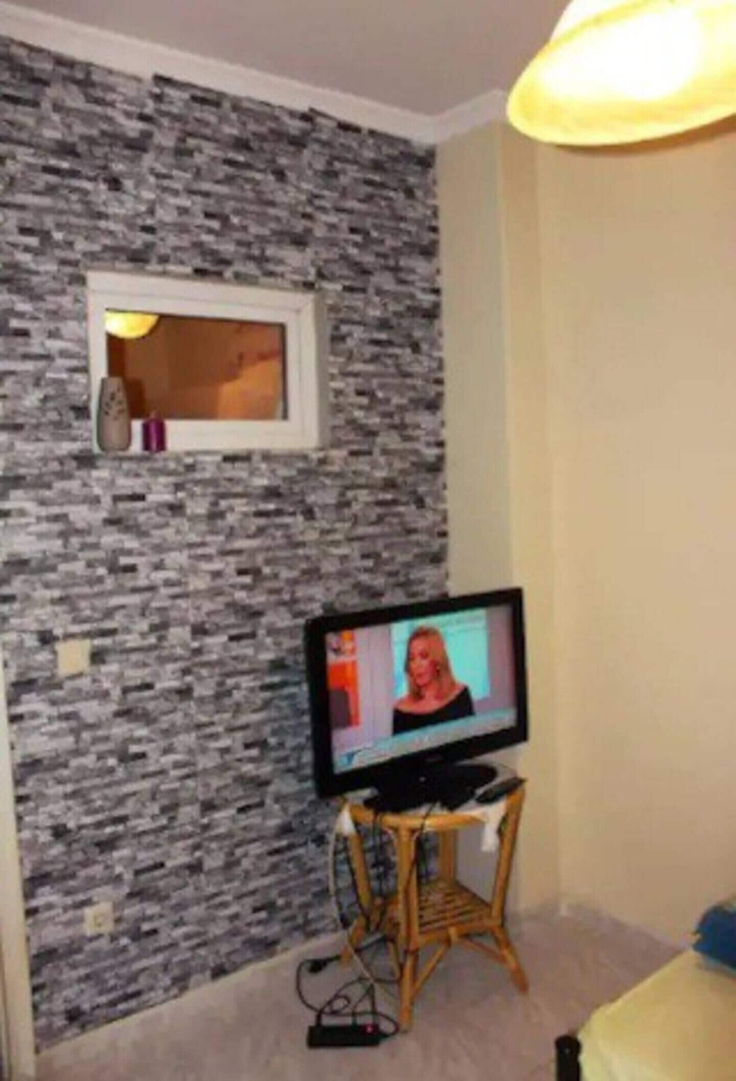 A room with a stone-patterned wall, a television on a stand, a small table, and a bed.
