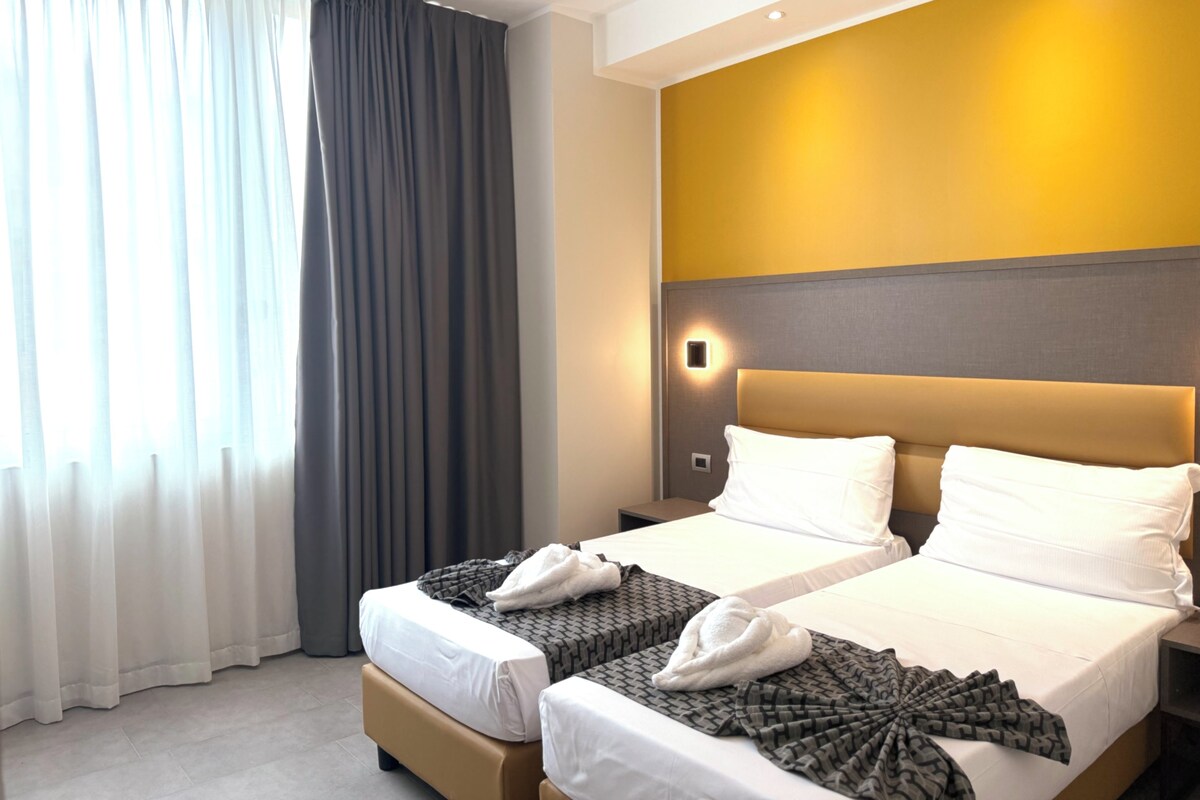 Hotellihuone, jossa on kaksi sänkyä, suuri ikkuna verhoilla, seinään kiinnitetty valaisin ja pelkistetty seinä, jossa on keltainen korosteväri.