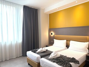 Hotellihuone, jossa on kaksi sänkyä, suuri ikkuna verhoilla, seinään kiinnitetty valaisin ja pelkistetty seinä, jossa on keltainen korosteväri.