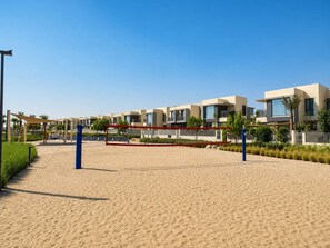 Ein sandiger Strand mit einem Volleyballnetz, moderne Gebäude im Hintergrund und einem klaren, blauen Himmel.