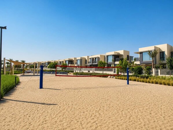 A sandy beach with a volleyball net, modern buildings in the background, and a clear blue sky. - Private Garden Villa | 3BR + Maid’s, Dubai Hills (Dubai)