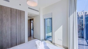 A modern hotel room with a large bed, a wooden panel wall, a large window with a view of high-rise buildings, and a door leading to a hallway. - Luxury 2BR Plus Maids, High-Floor Marina Views (Dubai)