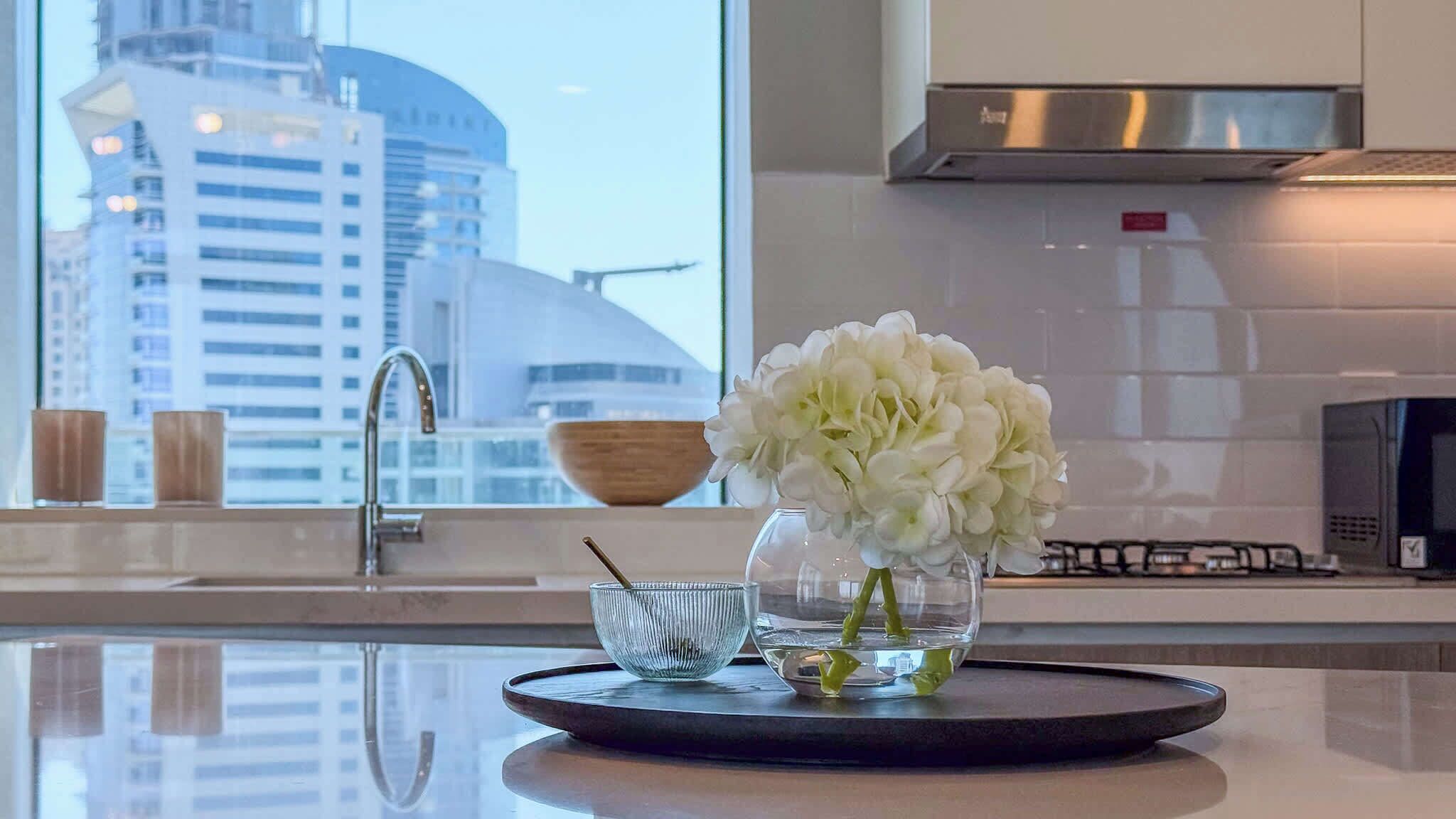 Uma cozinha moderna com um exaustor de aço inoxidável, um vaso de vidro com flores brancas sobre uma bandeja, e vista para o horizonte urbano pela janela.