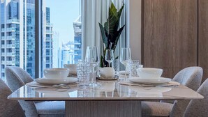 A dining table set with white plates, bowls, and glasses, surrounded by grey upholstered chairs, with a view of tall buildings in the background. - Luxury 2BR Plus Maids, High-Floor Marina Views (Dubai)
