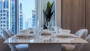 A dining table set with white plates, bowls, and glasses, surrounded by grey upholstered chairs, with a view of tall buildings in the background. - Luxury 2BR Plus Maids, High-Floor Marina Views (Dubai)