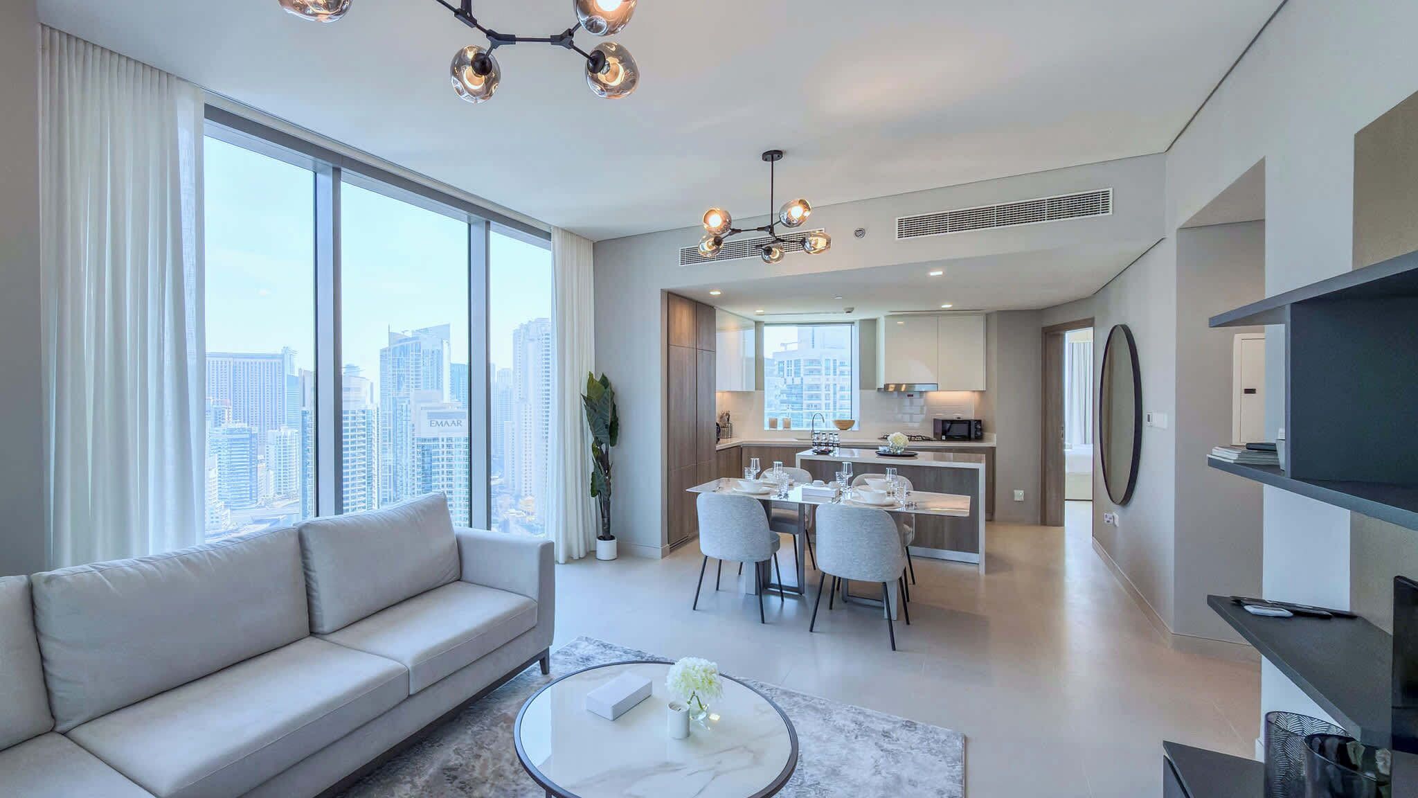 A modern living room with a sofa, dining table, and chairs, a kitchen area, and a large window with a city view.