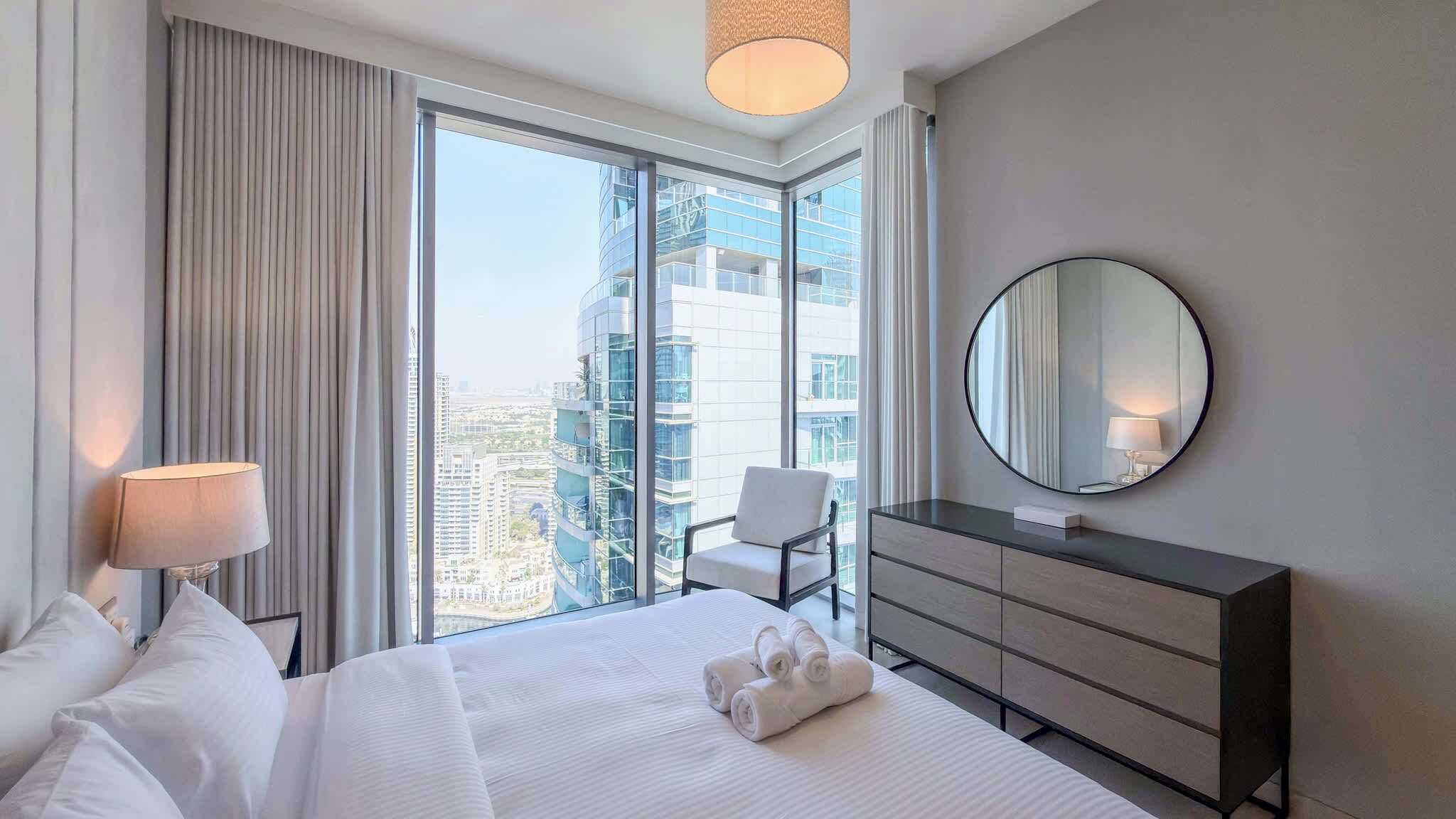 A hotel room with a large bed, a round mirror, a dresser, and a chair.