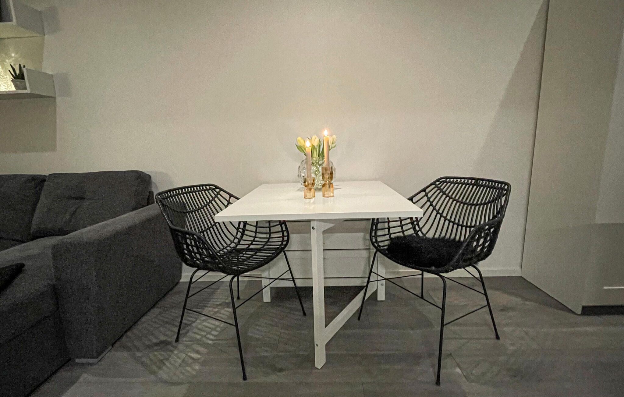 A small dining area with a white table and two black wireframe chairs. A vase with candles and yellow flowers is on the table.