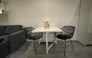 Ein kleiner Essbereich mit einem weißen Tisch und zwei schwarzen Stuhlgestellen. Auf dem Tisch steht eine Vase mit Kerzen und gelben Blumen.