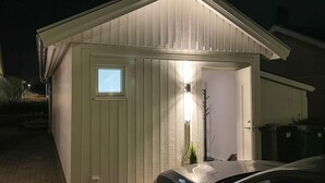 Ein kleines, eingeschossiges Gebäude mit einem geneigten Dach, einer Tür mit Fenster und einer Lampe an der Außenwand.