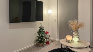 Ein modernes Wohnzimmer mit einem an der Wand befestigten Flachbildfernseher, einer Stehlampe, einem kleinen Weihnachtsbaum, einer Santa Claus Figur und einem runden Tisch mit dekorativen Gegenständen.
