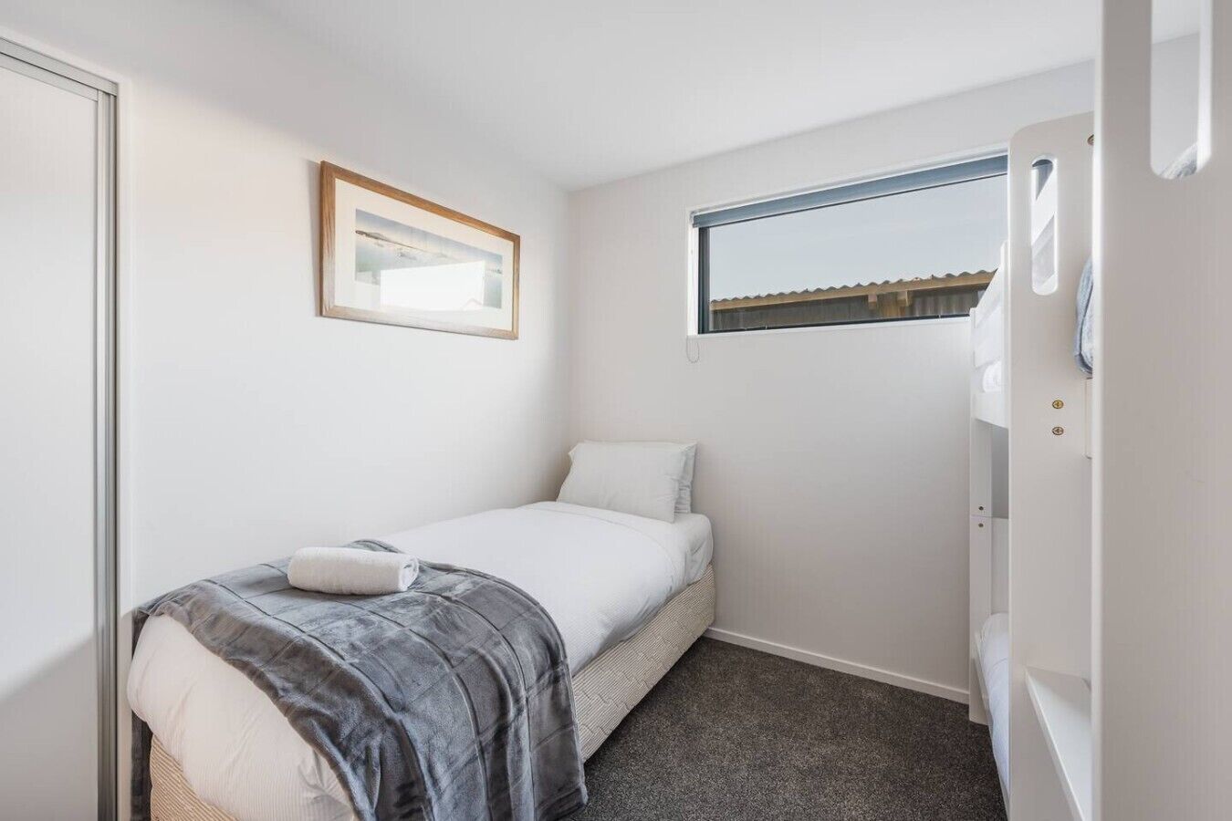 A small, single-bed room with a window, a framed picture on the wall, and a door to the right.