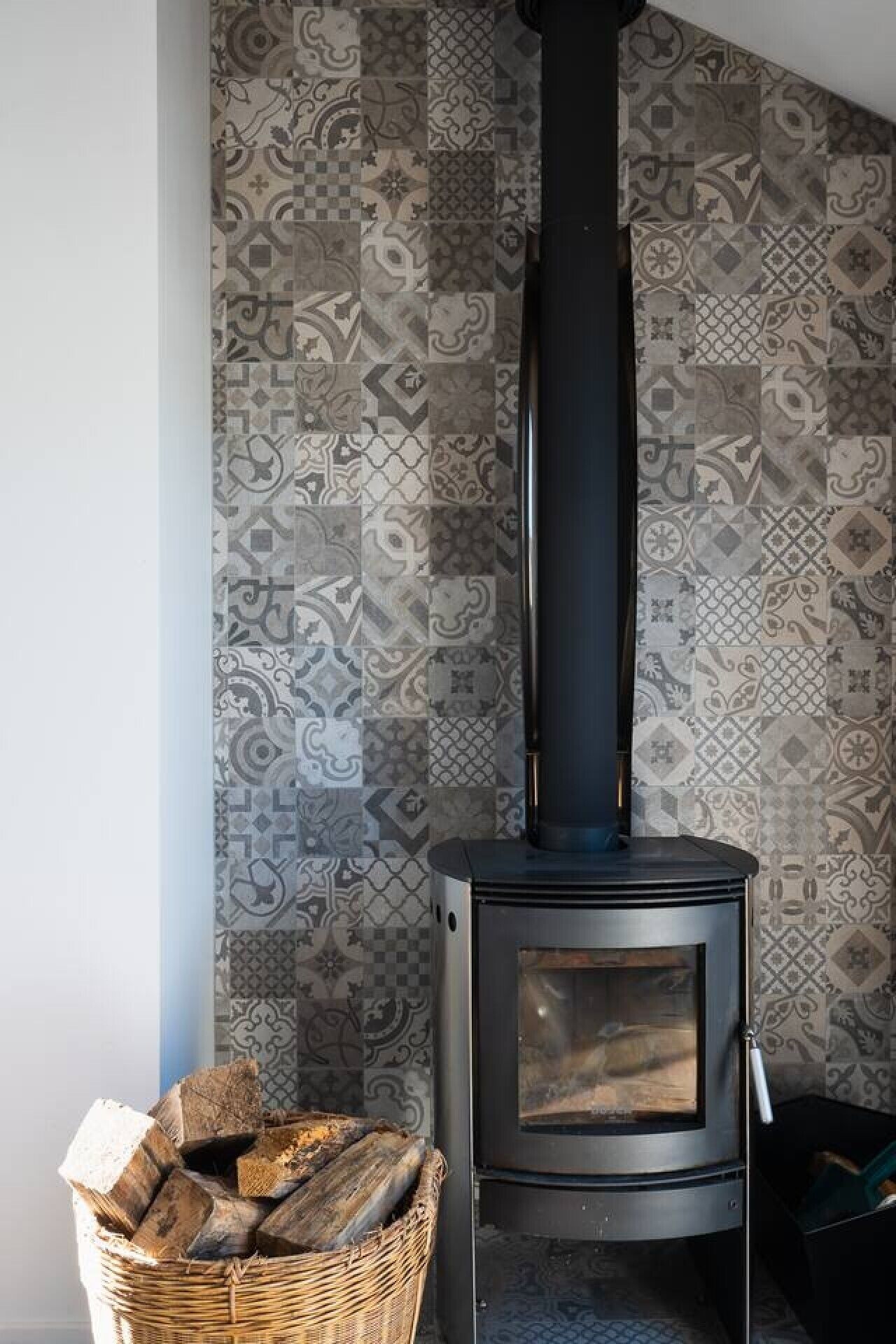 A wood-burning stove with a black chimney, a woven basket of firewood, and patterned wall tiles.