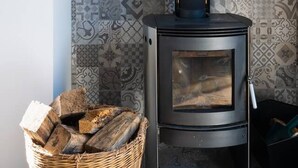 A wood-burning stove with a black chimney, a woven basket of firewood, and patterned wall tiles.