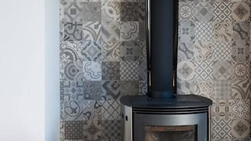 A wood-burning stove with a black chimney, a woven basket of firewood, and patterned wall tiles.