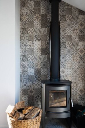 A wood-burning stove with a black chimney, a woven basket of firewood, and patterned wall tiles.