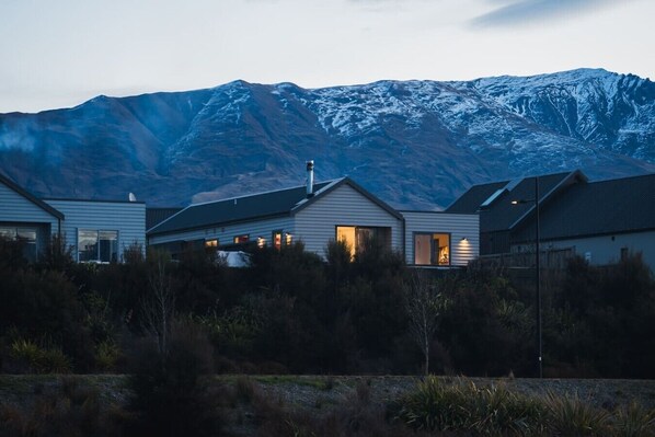 A modern house with a mountain backdrop, featuring a large window and a visible chimney.
