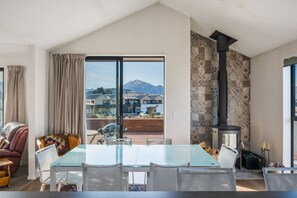 A modern living room with a glass dining table, comfortable seating, a large window with a mountain view, and a wood-burning stove.
