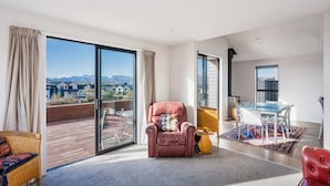 A spacious living area with a large sliding glass door leading to a balcony, a leather armchair, a dining table with chairs, and a view of a mountainous landscape.