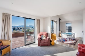 A spacious living area with a large sliding glass door leading to a balcony, a leather armchair, a dining table with chairs, and a view of a mountainous landscape.