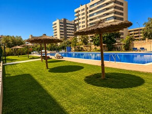 A swimming pool area with grass, umbrellas, and a building in the background. - Spacious 2BR Pool Parking Near the Beach (Alicante)